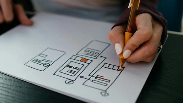 Una persona utiliza un bolígrafo naranja para dibujar diagramas de flujo o interfaces de usuario en papel.